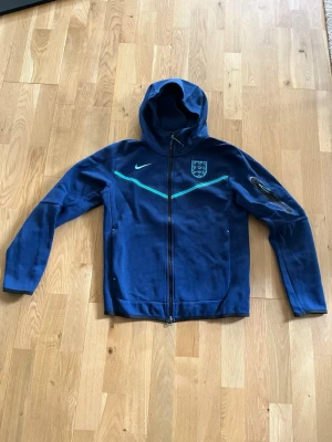 Blå England hoodie med dragkedja - Snygg blå hoodie från Nike med Englands landslagsmärke på bröstet och ljusblå detaljer över bröstet. Tröjan har dragkedja framtill, huva och långa ärmar. Perfekt för dig som gillar sportig stil och vill visa ditt stöd för England. Litet slitage på England emblemet.