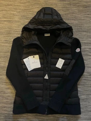 Svart pufferjacka med huva från Moncler - Snygg svart pufferjacka från Moncler med vadderad front och stickade ärmar. Jackan har en stor huva, dragkedja framtill och Moncler-logga på ärmen. Perfekt för kalla dagar och riktigt trendig look.