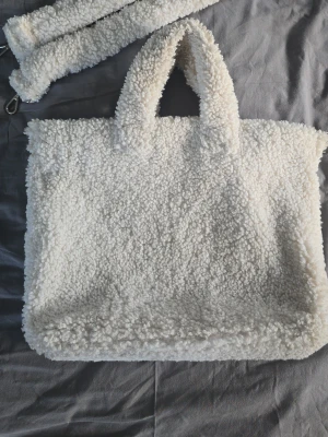 Vit fluffig shoppingväska i teddy - Supermjuk och fluffig shoppingväska i teddy, off-white färg, matchande handtag och axelrem. Väskan har en rektangulär form och är perfekt för dig som gillar mysiga accessoarer. Teddy-materialet ger en trendig och unik look. 