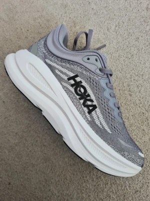 Grå Hoka sneakers med tjock sula - Snygga grå sneakers från Hoka med vit tjock sula och svart logga på sidan. Ovandelen är i meshmaterial för bra ventilation och skorna har klassisk snörning. Perfekta för dig som gillar sportig stil och vill ha riktigt bekväma skor.