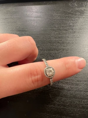 Silverfärgad ring med gnistrande stenar från Pandora - Snygg silverfärgad ring med en rund, stor klar sten i mitten omgiven av mindre gnistrande stenar. Ringen har även små stenar längs bandet vilket ger extra bling. Perfekt för dig som gillar stilrena men ändå iögonfallande smycken. Storlek 50