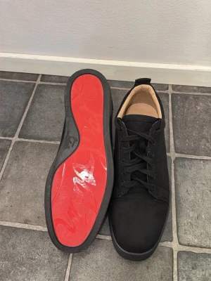 Svarta sneakers med röd sula - Stilrena svarta sneakers med ovandel i mockaliknande material och klassisk snörning. Insidan är beige och sulan är svart med en iögonfallande röd undersida. Perfekta för dig som vill ha en clean look med en twist.
