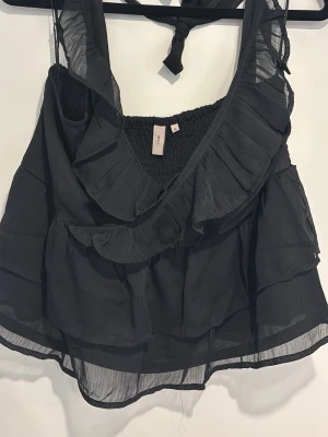 Volang topp från Nelly - Säljer en svart, ärmlös topp med volanger. Toppen har knytband i nacken skön passform där bak. Tunt, luftigt material som är perfekt till sommaren✨ 