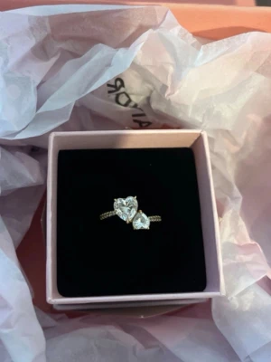 Pandora ring med hjärtformade stenar - Säljer en helt ny snygg guldpläterad ring från Pandora med två stora, gnistrande hjärtformade stenar. Ringen har en tvinnad ringband-design. Perfekt accessoar för dig som vill sticka ut lite extra. Säljer för att den inte passade. Asken och brev ingår.