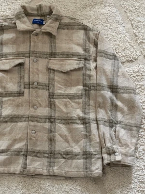 Beige overshirt med rutigt mönster - Snygg beige overshirt från Jack & Jones med rutigt mönster i grått. Två stora bröstfickor med lock och tryckknappar framtill.                                            XS sitter som S/M.   Oanvänd