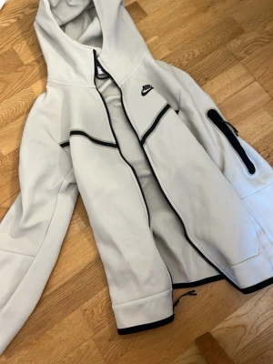 Vit Nike hoodie med svarta detaljer XS - Säljer en stilren vit hoodie från Nike i storlek XS. Hoodien har svarta detaljer längs dragkedjan och på ärmen, samt en praktisk huva. Tillverkad i mjukt material och har en ficka med dragkedja på ena ärmen. Perfekt för en sportig och avslappnad look.