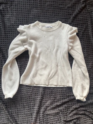 Vit ribbad tröja med volangärmar - Supersöt vit ribbad tröja med volangdetaljer vid axlarna och långa puffärmar. Tröjan har rund halsringning och är gjord i ett mjukt stickat material. Perfekt för dig som gillar en feminin och trendig stil.