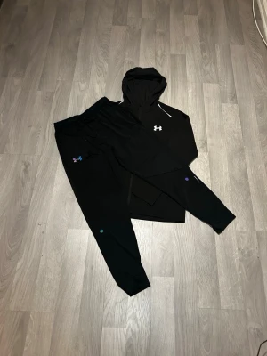 Svart träningsset från Under Armour - Snyggt svart träningsset från Under Armour med matchande hoodie och byxor. Hoodien har dragsko och logga på bröstet, medan byxorna har resår i midjan, dragsko och färgglad logga på benet. Perfekt för gymmet eller löprundan.