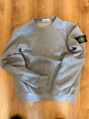 Grå sweatshirt från Stone Island - Snygg grå sweatshirt från Stone Island med klassisk rund hals och ribbade muddar. Tröjan har den ikoniska svarta logotypen med gult och grönt broderi på vänster ärm. Perfekt för en avslappnad och stilren look. Storlek S men kan även passa M. Knappt använd 