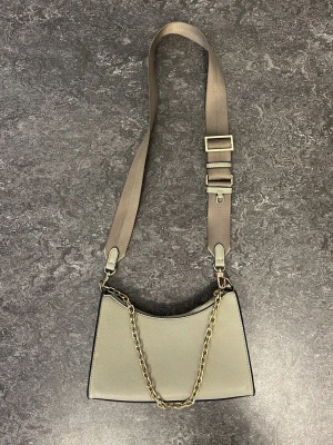 Beige axelremsväska med kedjedetalj - Stilren grön/beige handväska i slätt konstläder med justerbar bred axelrem i textil och snygg guldfärgad kedja som detalj. Väskan har minimalistisk design, perfekt för en trendig look.