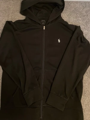 Svart hoodie från Polo Ralph Lauren - Snygg svart hoodie med dragkedja från Polo Ralph Lauren. Klassisk design med vit broderad logga på bröstet, huva och fickor framtill. Tillverkad i mjukt material som känns skönt mot huden. Perfekt för en chill och stilren look.