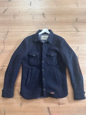 Mörkblå overshirt från Peak Performance - Snygg mörkblå overshirt från Peak Performance med två bröstfickor med lock och knappar, samt två sidofickor. Grov ullmix ger en robust känsla och förstärkta armbågar i kontrastmaterial. Perfekt lager på lager-plagg med klassisk krage och knappstängning.