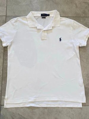 Vit pikétröja från Polo Ralph Lauren - Klassisk vit pikétröja från Polo Ralph Lauren i bomull med blå logga broderad på bröstet. Tröjan har krage, korta ärmar och knappar vid halsen. Perfekt till både jeans och shorts för en clean och tidlös look.