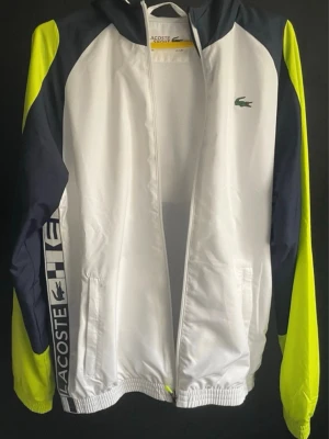 Lacoste vit och marinblå vindjacka - Snygg vindjacka från Lacoste i vitt, marinblått och neongult. Jackan har huva, dragkedja framtill, två fickor och elastiska muddar. Stora Lacoste-tryck på sidan och klassiska krokodilloggan på bröstet. Tillverkad i lätt och tåligt material, perfekt för en sportig look.