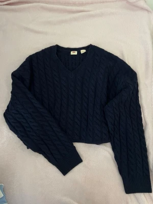 Mörkblå kabelstickad Levi's tröja - Snygg mörkblå kabelstickad tröja från Levi's med V-ringning och långa ärmar. Tröjan har ribbade muddar vid ärmslut och nederkant, samt ett klassiskt mönster som ger en tidlös look. Perfekt för dig som gillar stilrena och bekväma plagg. Aldrig använd.