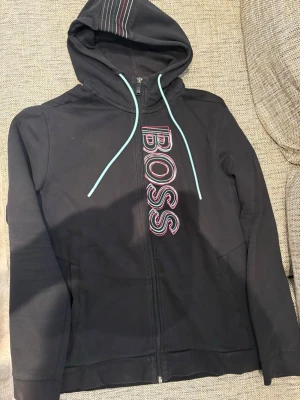 Svart hoodie med BOSS-tryck - Snygg svart hoodie med dragkedja och stor BOSS-logga i rosa och vitt på framsidan. Huvan har ljusblå snören och ett randigt mönster på toppen. Tillverkad i mjukt material, perfekt för en avslappnad look.