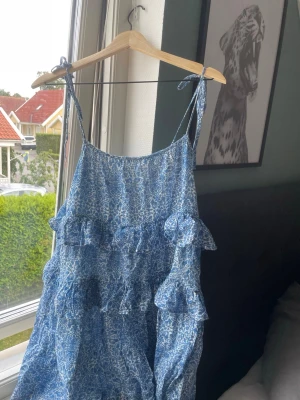 Blå blommig volangklänning - Söt blå klänning med vitt blommönster och volangdetaljer. Klänningen har smala axelband som knyts på axlarna och en luftig passform. Perfekt för varma dagar och ger en avslappnad men trendig look.
