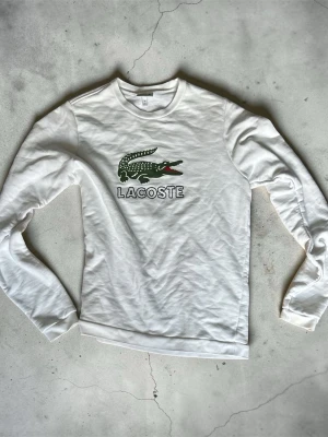 Vit långärmad tröja från Lacoste - Säljer en vit långärmad tröja från Lacoste med det klassiska gröna krokodilmotivet och tydlig logga på bröstet. Tröjan har rund halsringning och är gjord i mjukt material, perfekt för en clean och sportig look.