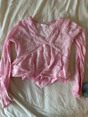 Rosa omlottblus från Gina Tricot - Söt rosa omlottblus från Gina Tricot i ett tunt, luftigt material. Blusen har långa ärmar med smock vid ärmsluten och ett v-format omlottyoke framtill. Perfekt för en trendig och avslappnad look.