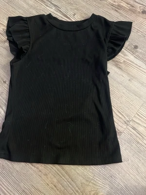 Svart ribbad topp med volangärm - Snygg svart ribbad topp med volangdetaljer på ärmarna. Toppen har rund halsringning och är tillverkad i ett mjukt, stretchigt material som sitter bekvämt. Perfekt för dig som gillar en enkel men trendig look.