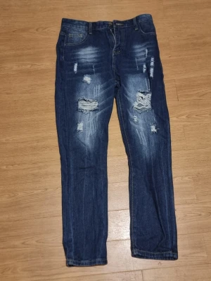 Mörkblåa jeans med hål  - INSTABOX!!! Säljer ett par blå jeans med trendiga slitningar och hål på framsidan. Jeansen har en klassisk femficksmodell och smal passform. Materialet är denim med en tvättad look och detaljerade slitningar för en cool streetstil.