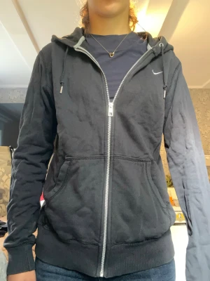 Svart zip-hoodie från Nike - Säljer en svart hoodie från Nike med dragkedja framtill, två fickor och justerbar huva med snören. Diskret Nike-logga på bröstet. Tillverkad i mjukt bomullsmaterial och har ribbade muddar vid ärmslut och nederkant. Finns ett litet hål i luvan, men det går inte igenom. 