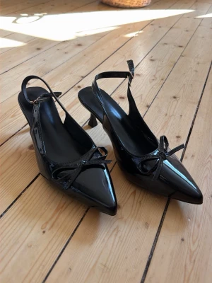 Svarta lackpumps med rosett - Säljer ett par snygga svarta pumps i lack med spetsig tå och liten klack. Skorna har en elegant rosettdetalj framtill och justerbar rem runt hälen. Perfekta för dig som vill ha en stilren och trendig look.