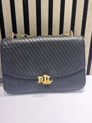 Svart axelväska med guldkedja RL - Pris kan diskuteras Snygg svart axelväska från Ralph Lauren med diagonal quiltning och guldfärgad kedjerem. Väskan har ett elegant RL-emblem i metall framtill och stilren rektangulär form. Perfekt för dig som gillar klassisk men trendig design.