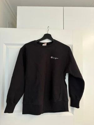 Svart sweatshirt från Champion - Klassisk svart sweatshirt från Champion med broderad logga på bröstet. Tröjan har rund halsringning, ribbade muddar vid ärmslut och nederkant samt är tillverkad i mjukt bomullsmaterial. 