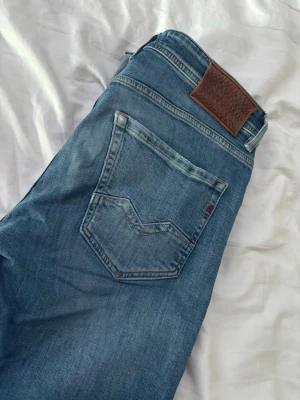 Blå jeans från Replay - Säljer ett par snygga blå jeans från Replay med klassisk femficksmodell och ljus tvätt. Jeansen har en skön passform och detaljer som läderpatch bak i midjan och Replay-logga på insidan. Perfekta till en avslappnad stil.