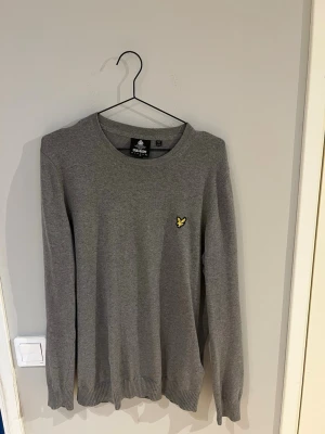 Grå tröja från Lyle & Scott - Snygg grå tröja från Lyle & Scott med rund halsringning och klassisk gul logga på bröstet. Tröjan har långa ärmar och ribbade muddar vid ärmslut och nederkant. Perfekt för en stilren och enkel look.