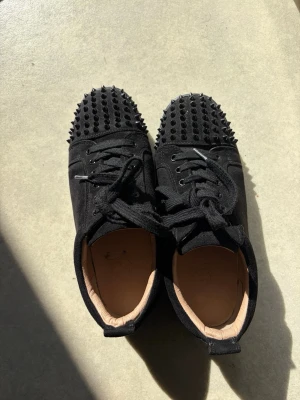 Svarta sneakers med nitar - Unika svarta sneakers från Christian Louboutin med svarta nitar på tån och klassisk röd sula. Skorna har snörning, svart mockaliknande material och beige innersula. Perfekta för dig som vill sticka ut med en edgy och lyxig stil. Säljer dessa då de är för stora för mig🙂