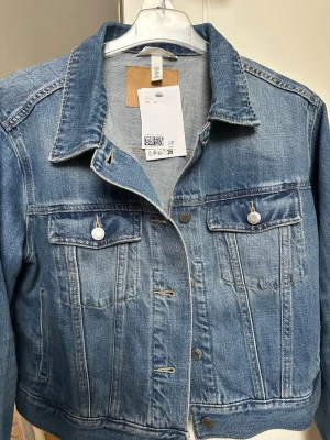 Klassisk blå jeansjacka i storlek M kort i modellen  - Snygg blå jeansjacka i klassisk modell med två bröstfickor med lock och knappar. Jackan har silverfärgade knappar framtill och på fickorna samt markerad krage. Perfekt att slänga över en hoodie eller t-shirt för en avslappnad look.