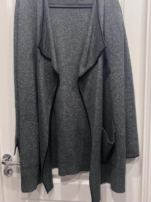 Grå öppen kofta från Zara Knit - Snygg gråmelerad öppen kofta från Zara Knit med svarta kanter och långa ärmar. Koftan har två fickor framtill och en avslappnad passform. Perfekt att slänga över en t-shirt eller topp för en chill look.