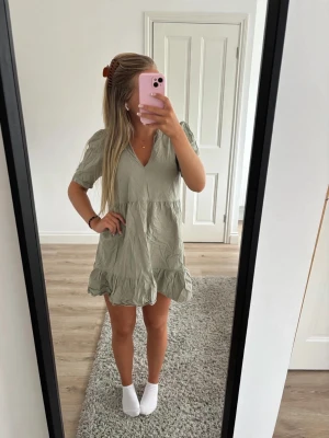 Ljusgrön kort klänning med volang - Supersöt ljusgrön kort klänning med v-ringning och volangdetaljer. Klänningen har korta ärmar och en lös, flowy passform som ger en avslappnad vibe. Perfekt för varma dagar och enkel att styla med smycken eller sneakers.