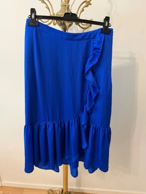 Kornblå volangkjol med omlott, 36 - Snygg kornblå kjol med volangdetaljer och omlott-design framtill. Kjolen har en längre modell och är tillverkad i ett lätt, flowy material som ger ett härligt fall. Perfekt för dig som vill sticka ut med färg och stil.