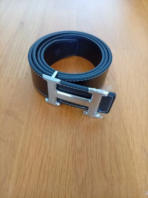Svart skärp med silverfärgat H-spänne Zara - Snyggt svart bälte med ett stort, silverfärgat H-format spänne. Bältet har vita kontrastsömmar längs kanterna och är gjort i slätt skinn. Perfekt för att lyfta vilken outfit som helst.