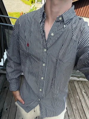 Randig skjorta från Ralph Lauren - Säljer en klassisk svart och vit randig skjorta från Ralph Lauren med broderad röd logga på bröstet. Skjortan har knappar framtill, lång ärm och button-down krage. Tillverkad i bomull och har en avslappnad classic fit.