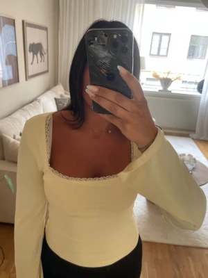 Ljusgul topp  med rosett i ryggen - Trendig ljusgul topp med långa ärmar och fyrkantig ringning. Toppen har fina spetsdetaljer vid halsen och en snygg omlottdesign med knyt i ryggen. Perfekt för dig som vill ha en feminin och stilren look.