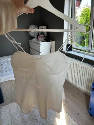 Vit satinlinnetopp med tunna band - Säljer en söt vit linnetopp i satin med tunna axelband som knyts över axlarna. Toppen har en lätt utsvängd passform och markerade kupor framtill. Baksidan är smockad för extra komfort och passform. Perfekt för sommarens alla tillfällen!