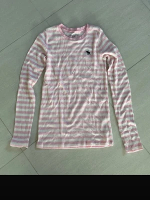 Rosa och vit randig tröja Abercrombie - Supersöt långärmad tröja från Abercrombie Kids i ribbad kvalitet. Tröjan har rosa och vita horisontella ränder, rund halsringning och en liten logga broderad på bröstet. Perfekt för en chill och avslappnad stil.