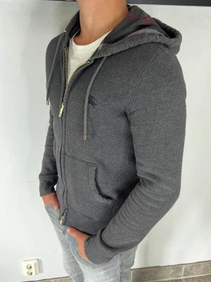 Burberry zip hoodie - Snygg mörkgrå hoodie med dragkedja framtill och två stora fickor. Tröjan har justerbar huva med snören och ribbade muddar vid ärmslut och nederkant. Perfekt för en avslappnad och stilren look. Materialet känns mjukt och bekvämt.