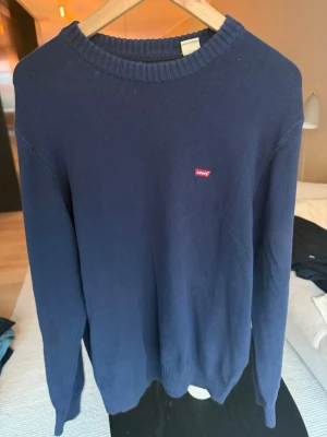 Mörkblå stickad tröja från Levi's - Mörkblå stickad tröja från Levi's med rund halsringning och diskret röd logotyp på bröstet. Tröjan har ribbstickade muddar vid ärmslut och nederkant. Perfekt för lager-på-lager och enkel att matcha med jeans.