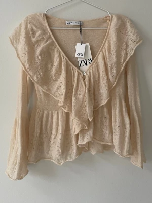 Stickad beige volangblus från Zara - Supersöt stickad beige blus från Zara med långa ärmar och stora volangdetaljer längs den djupa v-ringningen. 