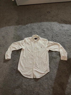 Vit skjorta från Ralph Lauren - Klassisk vit skjorta från Ralph Lauren med knappslå och broderad logga på bröstet. Skjortan har lång ärm, krage med knappar och är tillverkad i bomull. Perfekt för en clean och stilren look.