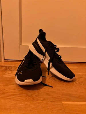Svarta Puma idrott sneakers med vit sula - Säljer ett par svarta idrott sneakers från Puma med vit sula och vit logga på sidan. Skorna har en sportig design med lågt skaft, snörning och dragflik bak. Ovandelen är i textil och syntetmaterial för en lätt känsla.