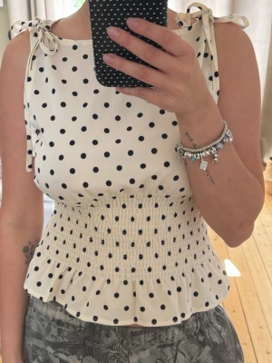 Prickig vit topp med knytband - Supersöt vit topp med svarta prickar och knytband på axlarna. Smockad midja och volang nertill ger en feminin och trendig look. Perfekt till jeans eller kjol för en fräsch stil.