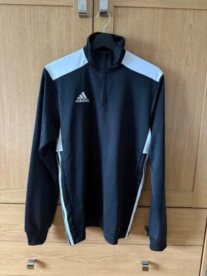 Svart träningströja från Adidas - Snygg svart träningströja från Adidas med vita detaljer på axlar och sidor. Jackan har halv dragkedja framtill, hög krage och långa ärmar med muddar. Perfekt för träning eller uppvärmning. 