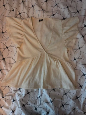 Krämvit topp från Gina Tricot - Säljer en krämvit topp från Gina Tricot i storlek XS. Toppen har v-ringning, korta volangärmar och en lätt utsvängd midja. Materialet är ribbat och ger en mjuk känsla. Perfekt att styla med jeans eller kjol!
