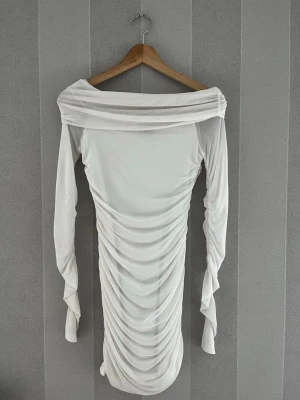 Vit offshoulder omlottklänning - Säljer en vit kortklänning med offshoulder och långa ärmar. Klänningen har snygga draperingar längs hela framsidan och baksidan, vilket ger en cool omlott-effekt. Perfekt för dig som vill sticka ut med en elegant och trendig look.